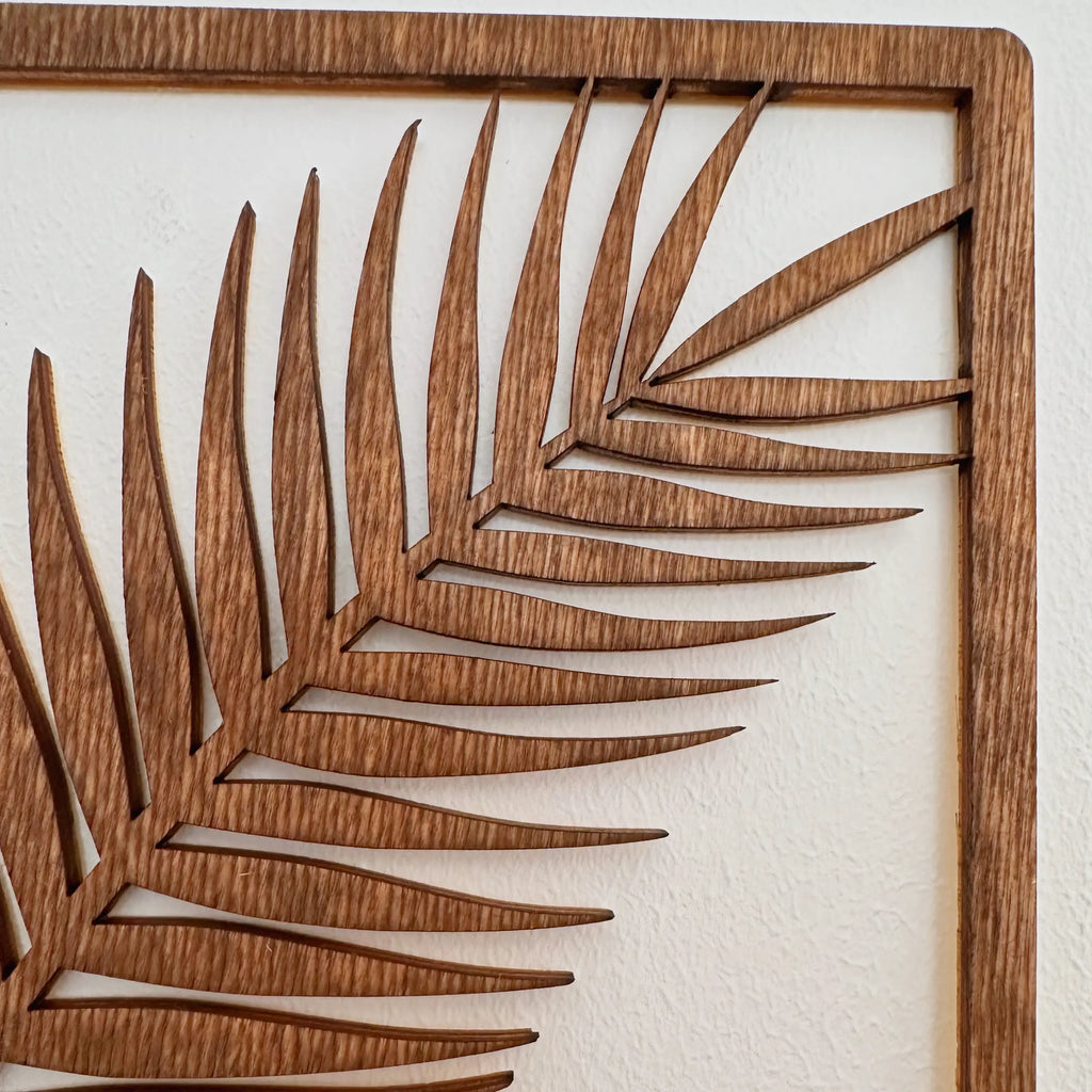 Vue rapprochée d'un tableau en bois qui symbolise une feuille de palmier, très graphique, sur mur blanc.